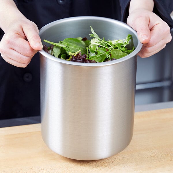 A person holding a Vollrath stainless steel pot of green salad on a table.