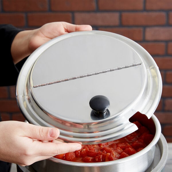 A person holding a Vollrath stainless steel lid over a pot of food.