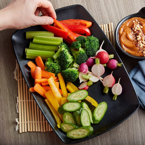 A hand holding a black rectangular platter of broccoli, carrots, and dip.