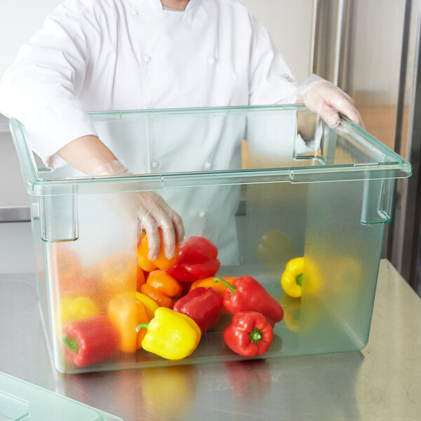 A person in a white coat and gloves putting red and yellow peppers into a Carlisle StorPlus green food storage box.