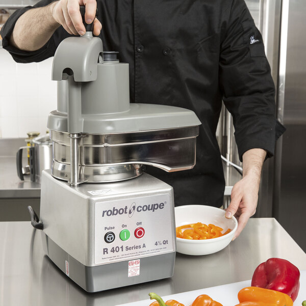 A man in a black coat using a Robot Coupe commercial food processor with a stainless steel bowl to chop peppers.