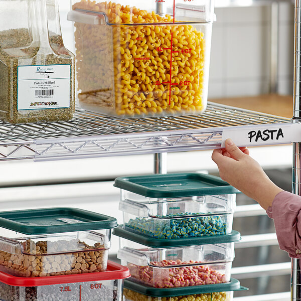 A hand holding a plastic label holder with a label for a container of pasta on a shelf.