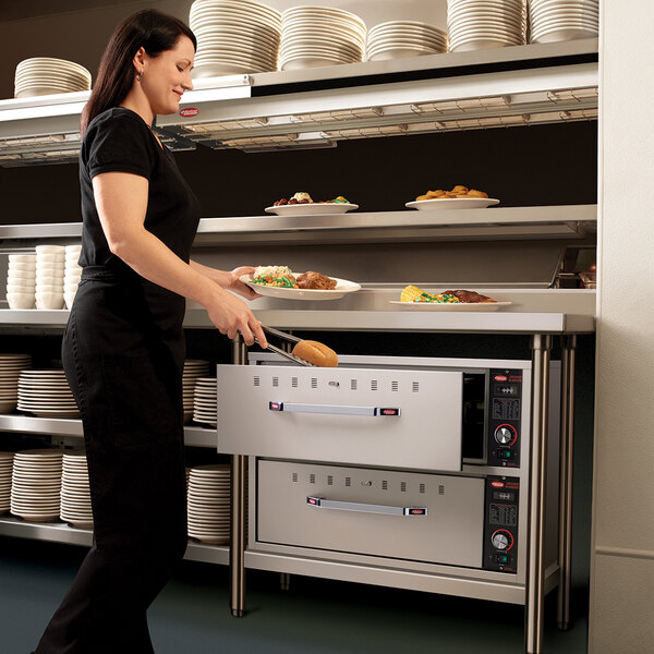 A woman in a black dress using a Hatco freestanding drawer warmer in a professional kitchen.