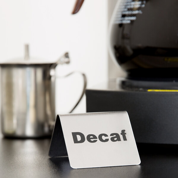 A Tablecraft stainless steel "Decaf" tent sign on a counter near a coffee pot.