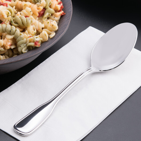A Oneida Stanford stainless steel serving spoon on a napkin next to a bowl of pasta.