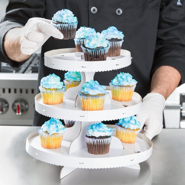 A person holding a Wilton white three tiered cupcake stand with cupcakes.