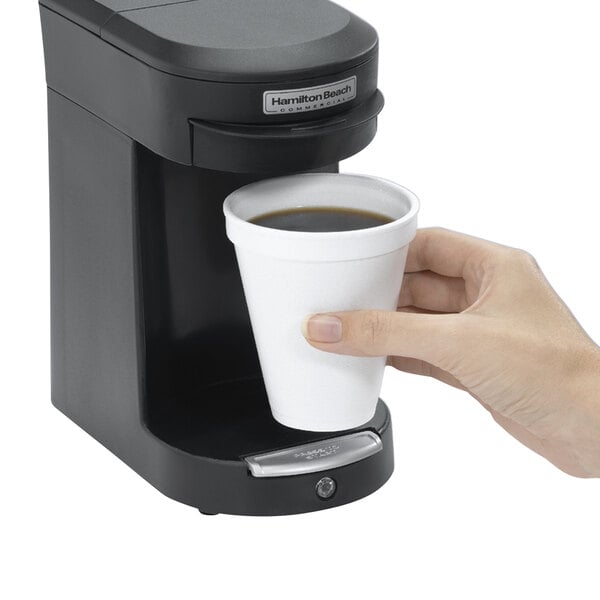 A hand using a Hamilton Beach black single serving pod coffee maker to fill a cup with coffee.