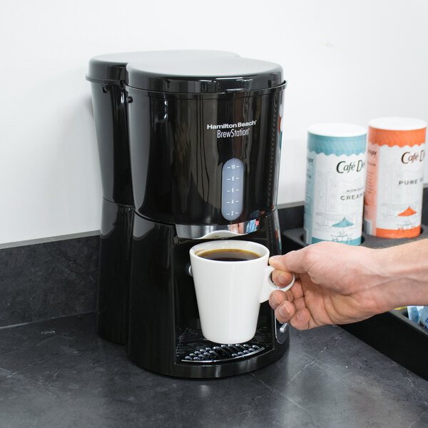 A person holding a white mug of coffee in front of a Hamilton Beach BrewStation coffee maker.