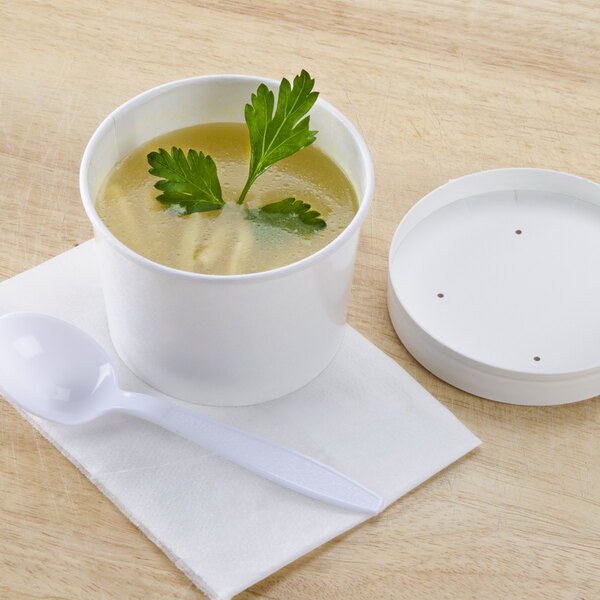 A white Huhtamaki poly paper food cup filled with soup and a plastic lid with a spoon on the table.