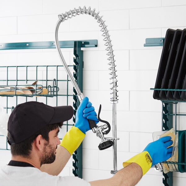 A man wearing blue gloves and yellow gloves uses a Regency pre-rinse spray valve and flex hose to clean a shelf in a professional kitchen.
