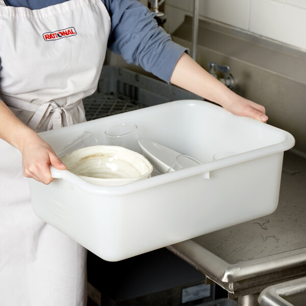 A woman in an apron holding a white Tablecraft plastic tub with dirty dishes inside.