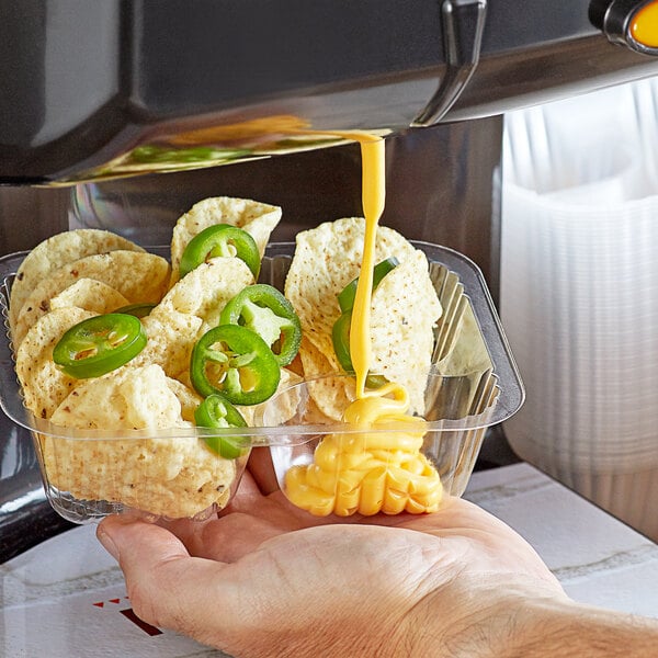 A hand holding a plastic container of chips with Muy Fresco Jalapeno Cheese Sauce and jalapeno slices.
