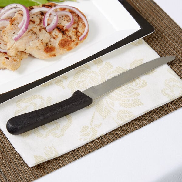 An American Metalcraft stainless steel steak knife with a black plastic handle on a napkin next to a plate of food.