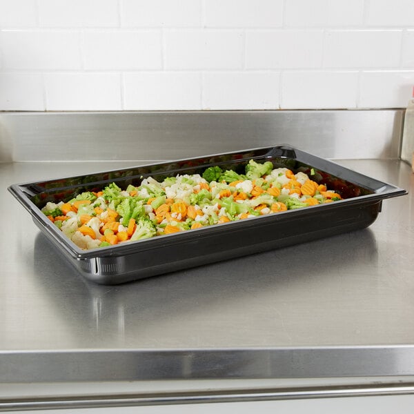 A Vollrath black plastic food pan on a counter with a tray of vegetables in it.