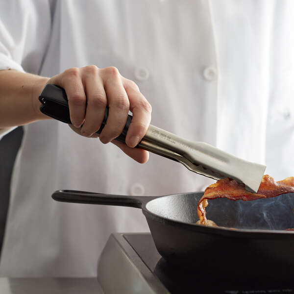 A hand with a Vollrath VersaGrip tong with a black handle holding bacon over a skillet.