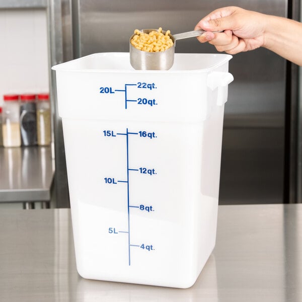 A hand using a measuring cup to pour macaroni into a white Cambro food container.