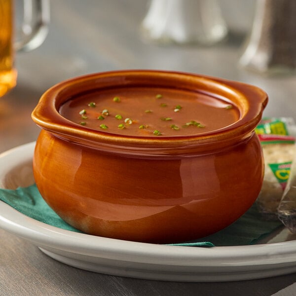An Acopa brown stoneware onion soup bowl filled with soup on a plate on a restaurant table.