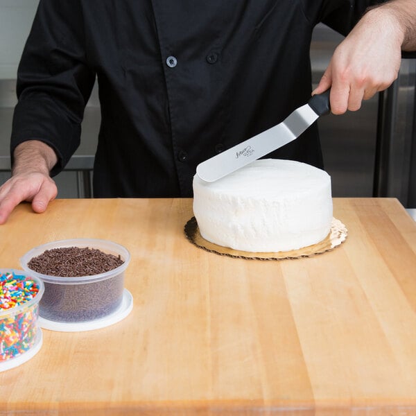 A person using an Ateco offset spatula to cut a white cake with chocolate sprinkles.