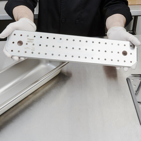 A person in gloves holding a Vollrath stainless steel tray with a metal piece in the bottom.