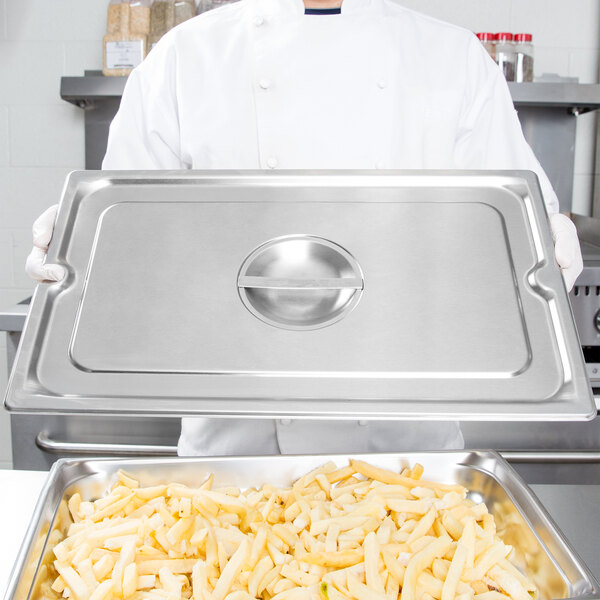 A chef holding a Vollrath stainless steel slotted cover over a tray of french fries.
