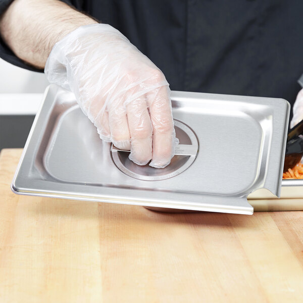 A person wearing plastic gloves holding a Vollrath stainless steel slotted cover over a tray of food.