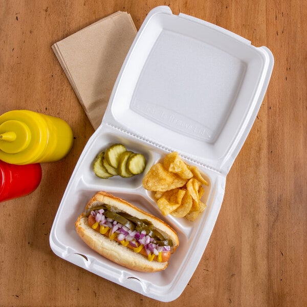 A Dart white foam takeout container with a hot dog, mustard, pickles, and chips.