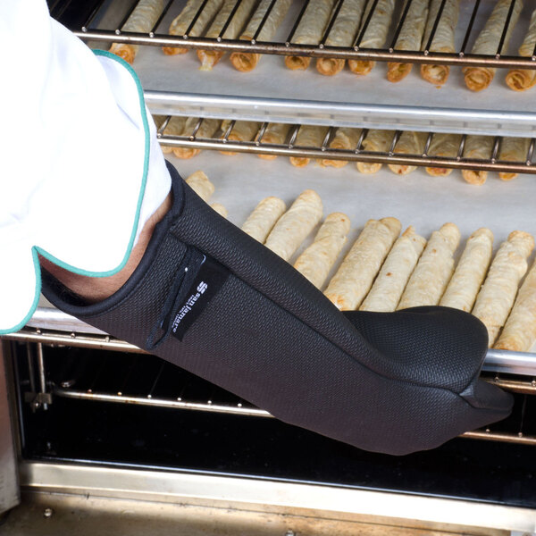 A person wearing a San Jamar black UltiGrips puppet style oven mitt holding a tray of pastries on a counter.
