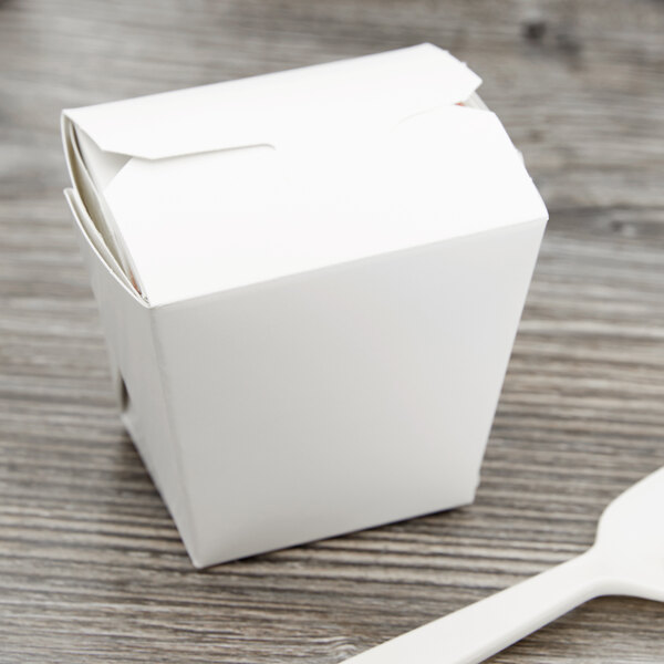 A white Fold-Pak take-out container with a lid and a plastic spoon on a wooden table.