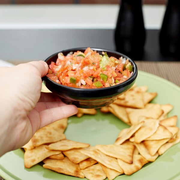 A hand holding a Carlisle black salsa bowl full of salsa and chips.