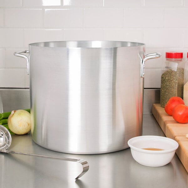 A large silver Vollrath Arkadia stock pot with handles next to a bowl of vegetables.