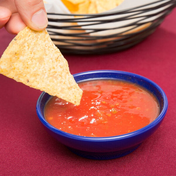 A hand holding a chip dipping into a Carlisle cobalt blue salsa dish filled with salsa.
