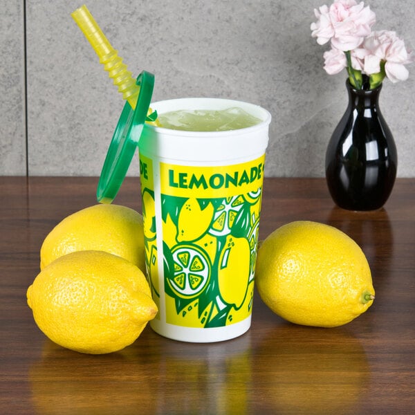 A yellow and green plastic lemonade cup with a white lid and a straw next to lemons.