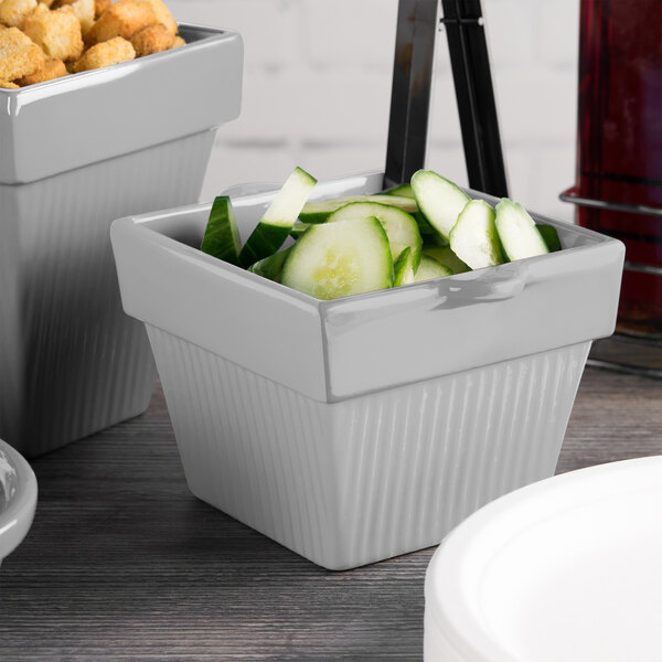 A white Tablecraft square condiment bowl with cucumbers and crackers on a table.