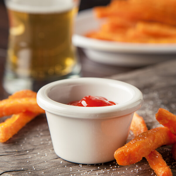 A bowl of Carlisle bone white ramekin filled with ketchup next to a bowl of french fries on a table.