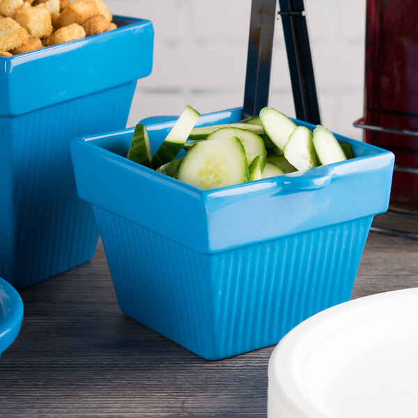 A Tablecraft sky blue square condiment bowl with cucumbers inside, on a table next to a white container.