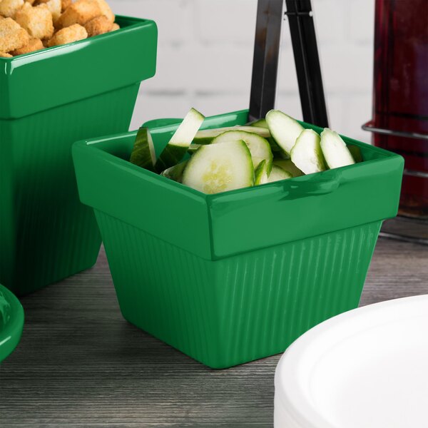 A green Tablecraft square condiment bowl with cucumbers in it on a counter.