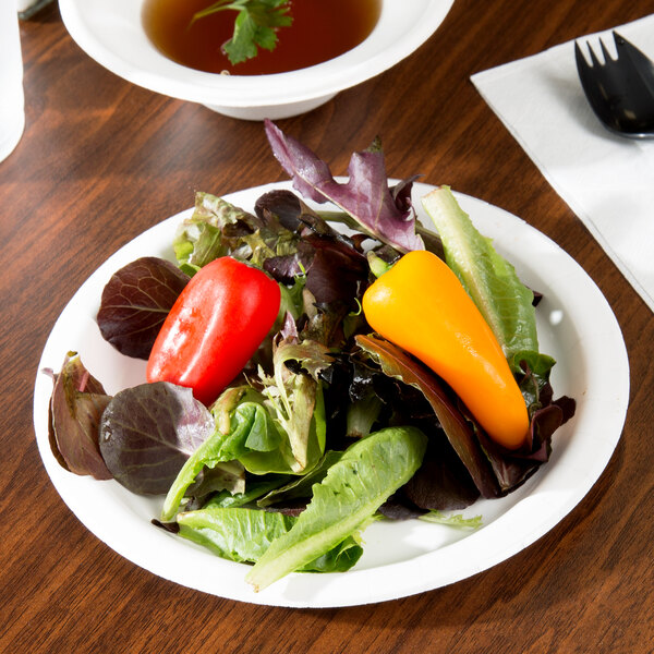 A white paper plate with a salad, small pepper, and bowl of soup on a counter.