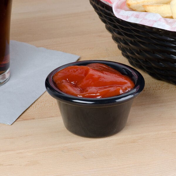 A black Carlisle smooth plastic ramekin filled with ketchup next to a basket of french fries.