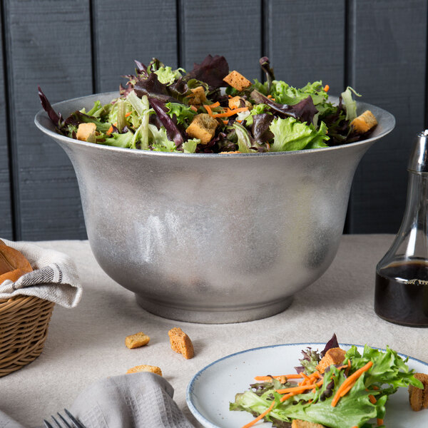 A Tablecraft natural cast aluminum tulip salad bowl on a table with a plate of salad and croutons.