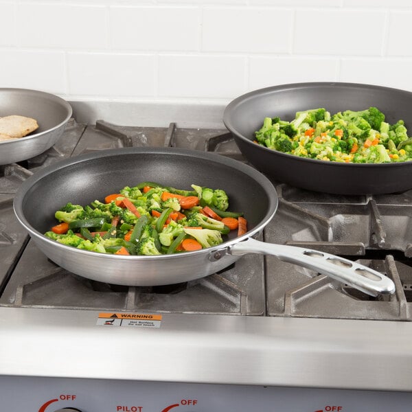 Two Vollrath Wear-Ever aluminum non-stick fry pans on a stove with vegetables cooking in one.