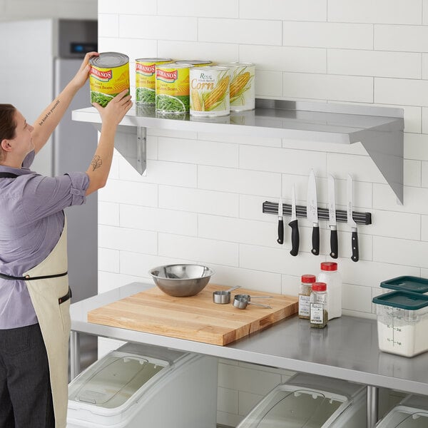 A woman in a professional kitchen using a Regency stainless steel wall shelf to put food in a white container with a clear lid.