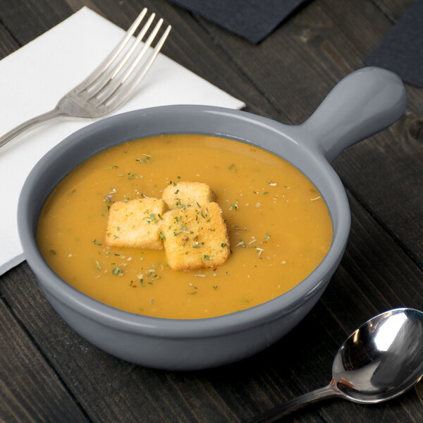 A gray Tablecraft cast aluminum soup bowl with a handle filled with soup and croutons with a spoon next to it.