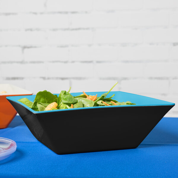 A blue and black square melamine bowl with salad on a table.