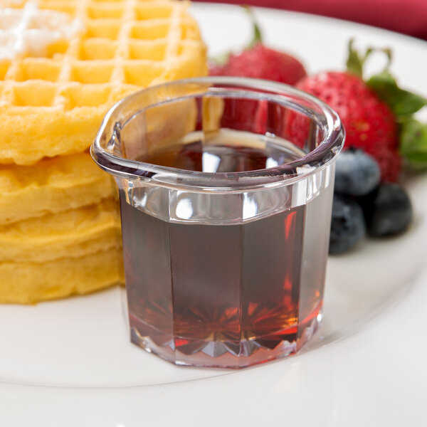 A clear plastic cup filled with syrup next to waffles and berries.