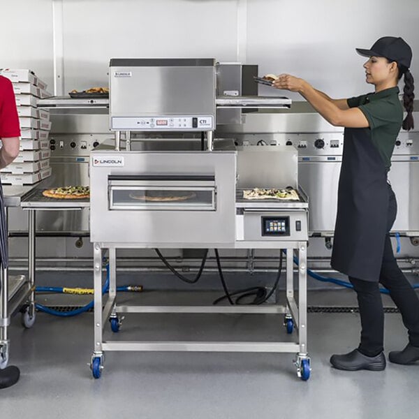 A woman and a man using a Lincoln Electric Conveyor Oven to cook a pizza.