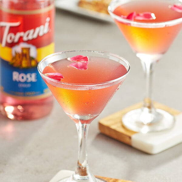 A glass of pink liquid with rose petals in it, with a Torani Rose Flavoring Syrup bottle on the table.