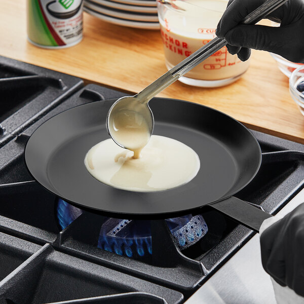 A person using a Matfer Bourgeat black carbon steel crepe pan to pour liquid into a pan on a stove.