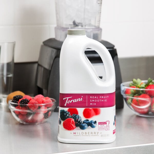 A jug of Torani Wildberry Fruit Smoothie Mix on a counter with bowls of strawberries, raspberries, and blackberries.