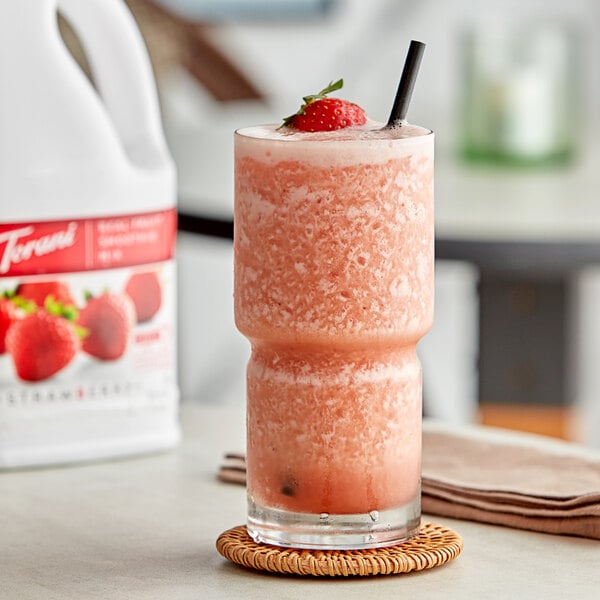 A glass of pink strawberry fruit smoothie with strawberries on the rim next to a bottle of Torani Strawberry Fruit Smoothie Mix.