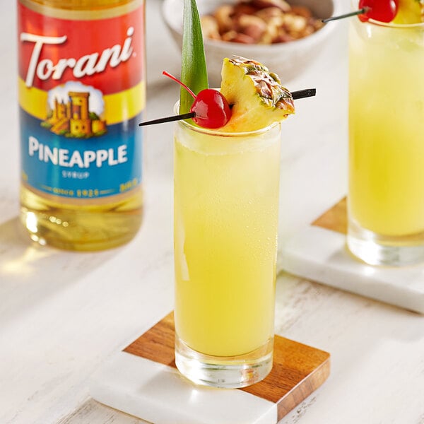 A glass of yellow liquid with a pineapple and a cherry on the rim.